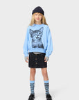 Child wearing a light blue sweatshirt with a cat print, black skirt, and striped socks on a white background