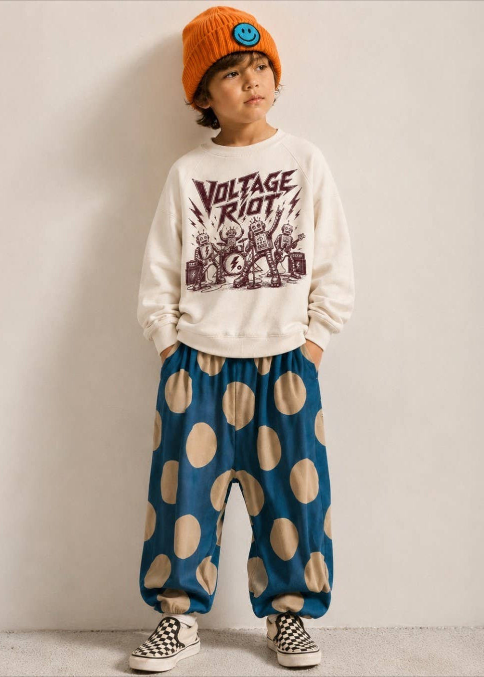 A child model wearing a cream crewneck sweatshirt with a dark graphic print, blue pants with large beige polka dots, a bright orange beanie, and black-and-white checkered sneakers, standing against a plain light gray wall.