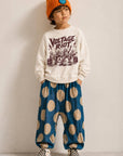 A child model wearing a cream crewneck sweatshirt with a dark graphic print, blue pants with large beige polka dots, a bright orange beanie, and black-and-white checkered sneakers, standing against a plain light gray wall.