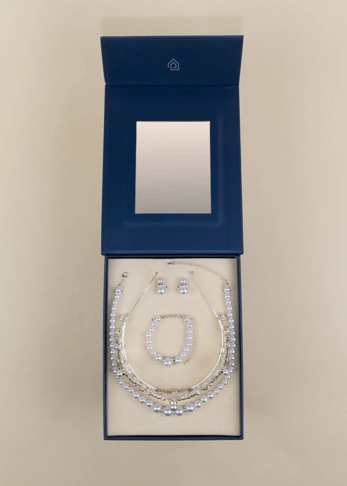 A jewelry set displayed inside an open dark blue presentation box with a mirror on the inside lid. The set includes a silver-toned necklace, bracelet, and earrings with clear rhinestone-like stones, arranged on a cream insert against a light beige background.