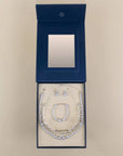 A jewelry set displayed inside an open dark blue presentation box with a mirror on the inside lid. The set includes a silver-toned necklace, bracelet, and earrings with clear rhinestone-like stones, arranged on a cream insert against a light beige background.