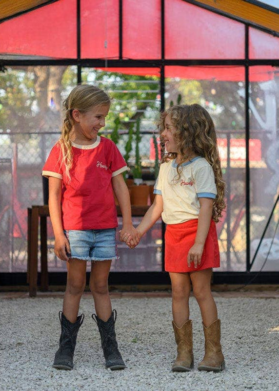 Two children holding hands in a greenhouse with plants and a colorful tent-like structure in the background.