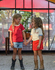 Two children holding hands in a greenhouse with plants and a colorful tent-like structure in the background.