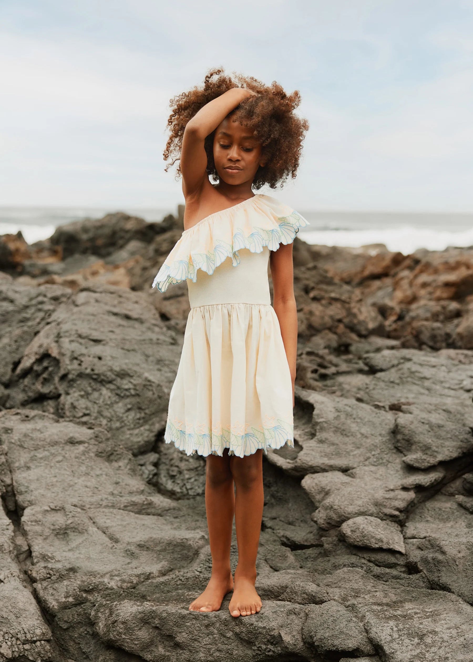 Young girl in a white dress standing on rocky terrain with a coastal background