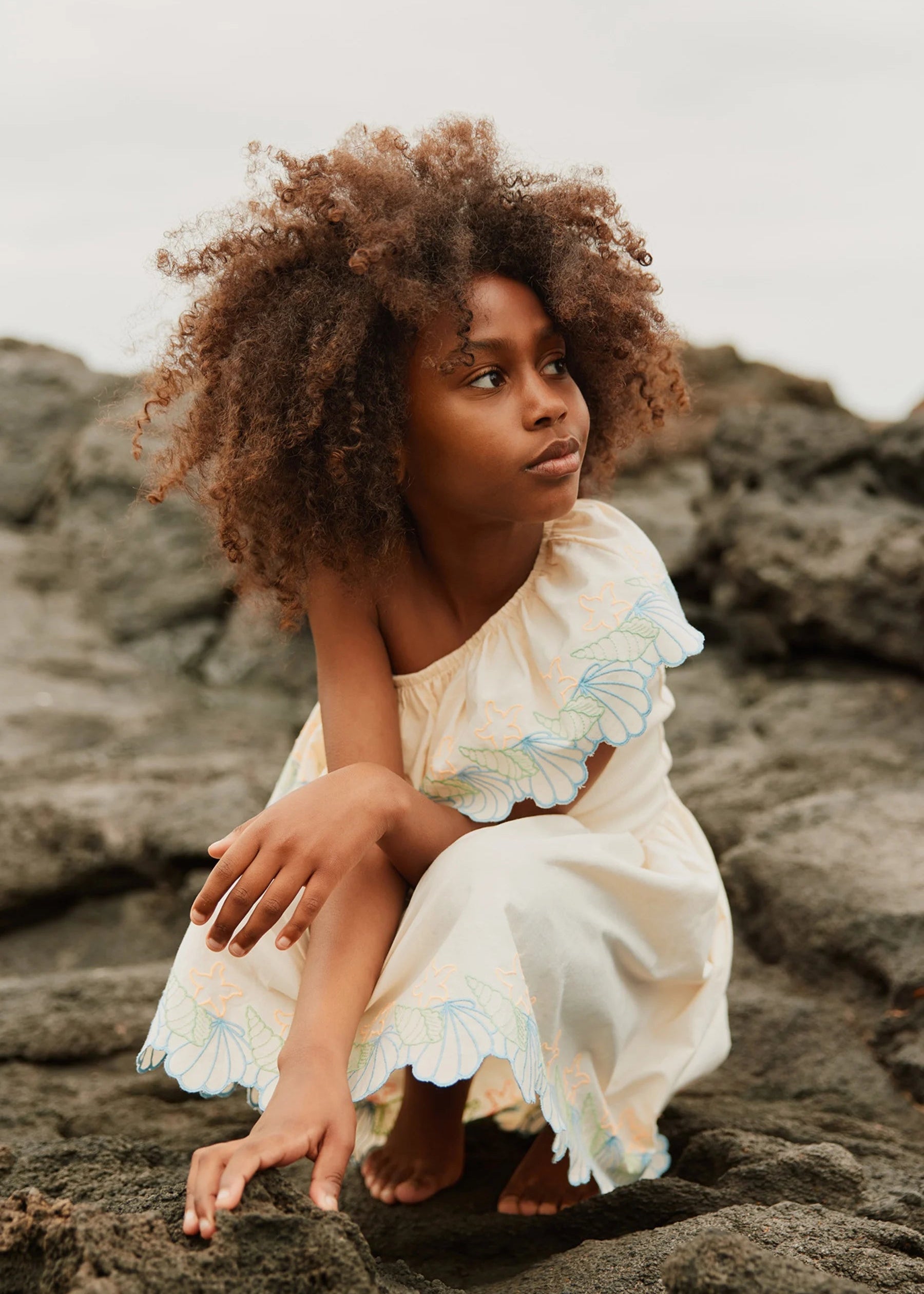 Young girl in a white dress with floral details sitting on rocks.