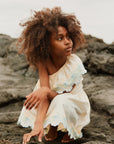 Young girl in a white dress with floral details sitting on rocks.
