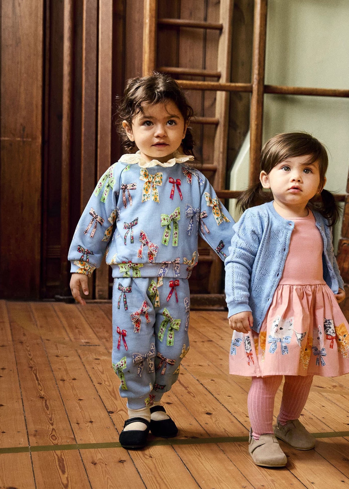 Two children in matching outfits standing on a wooden floor with wooden crates in the background.