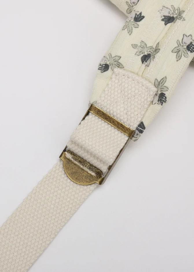 Close-up of a cream-colored woven strap with a delicate gray and black floral pattern, featuring a brass buckle.
