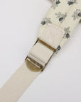 Close-up of a cream-colored woven strap with a delicate gray and black floral pattern, featuring a brass buckle.