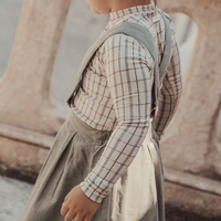 Child wearing a plaid shirt and gray dress standing in front of a stone wall.
