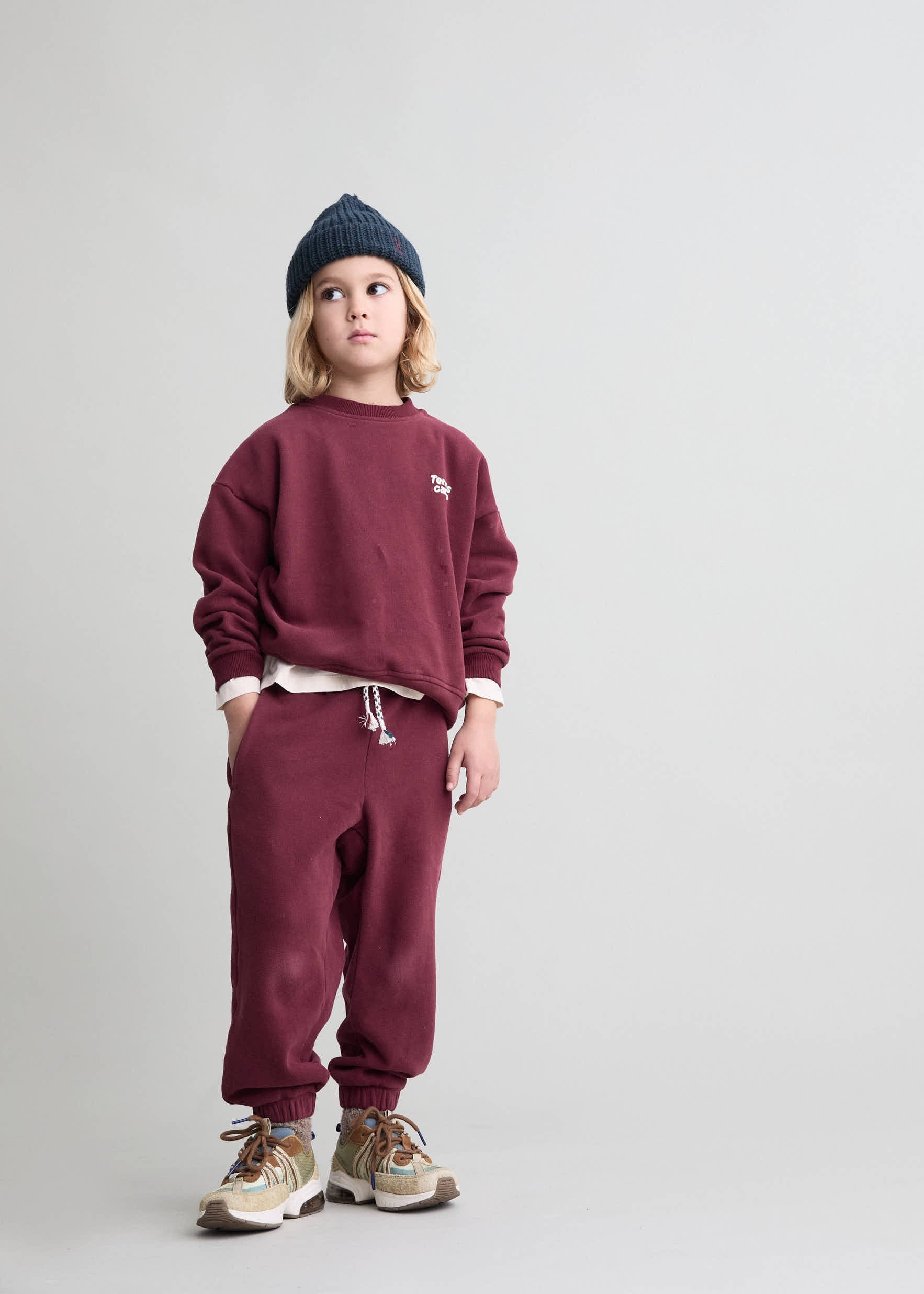 Child wearing a maroon sweatshirt and pants with a beanie on a light gray background