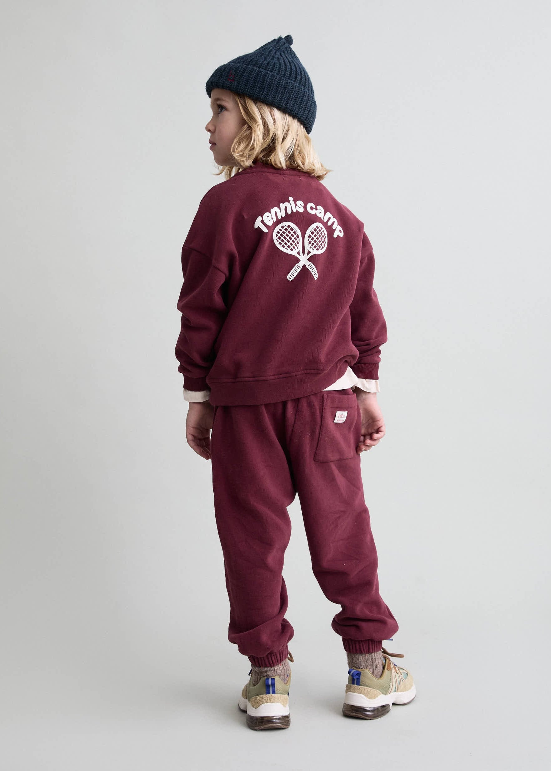 Child wearing a maroon tracksuit with 'Tennis Camp' text on the back against a plain background