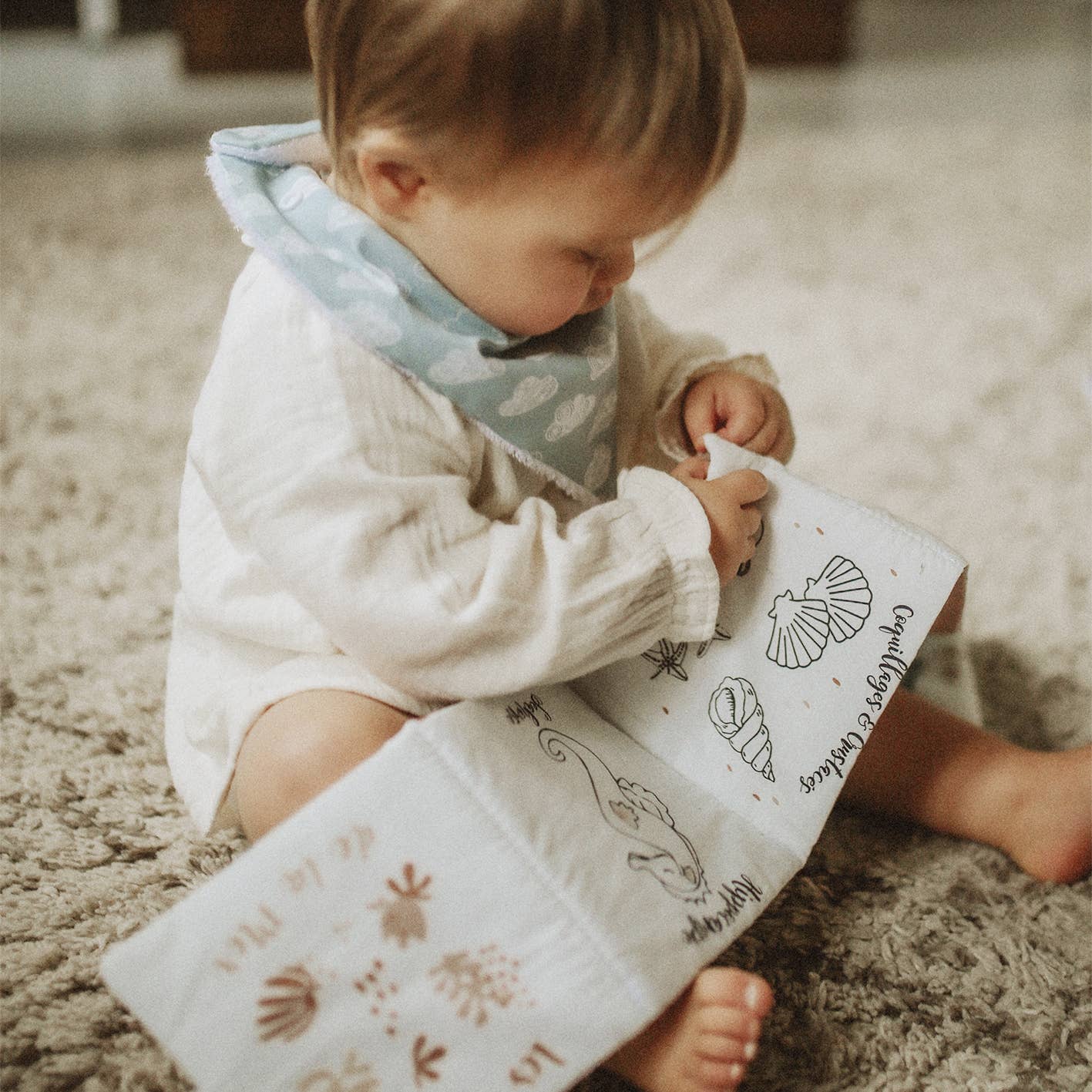 A baby sitting on a textured beige rug, holding a white cloth book with black illustrations and brown botanical designs. The baby is wearing a white long-sleeve bodysuit and a light blue patterned bib.