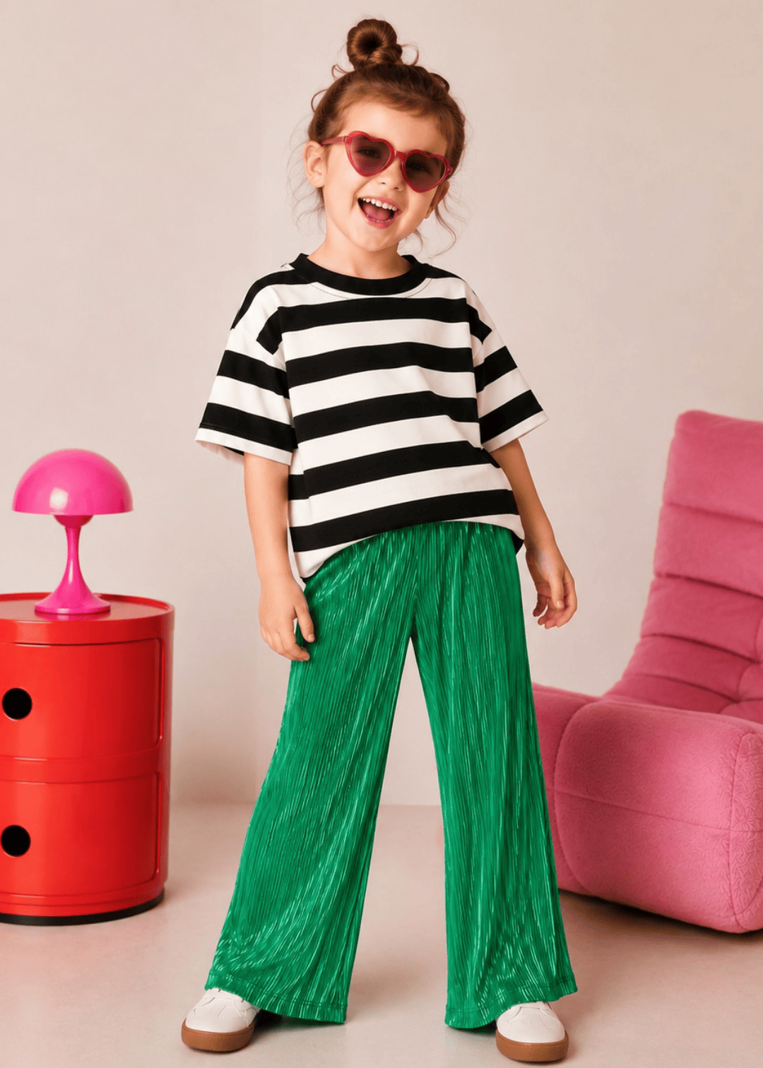 Child wearing a black and white striped shirt and green pants standing in a room with a red side table and pink chair.