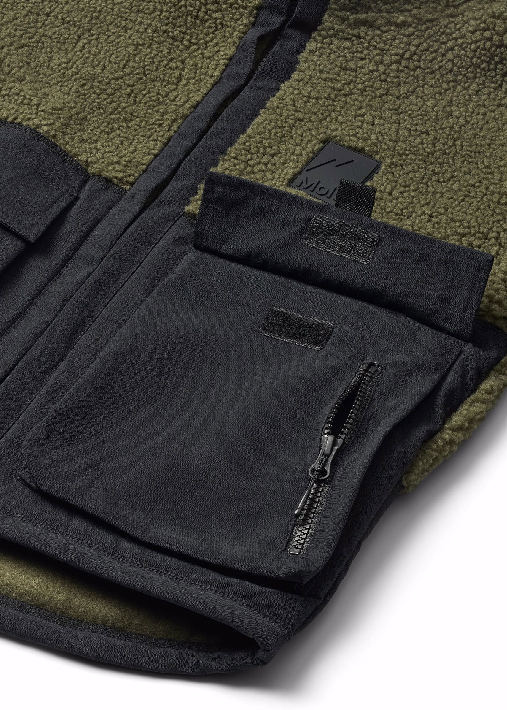 Close-up of a fleece-lined jacket showing a black front zip pocket with a zipper and a small black brand tab, alongside an olive green sherpa lining.