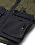 Close-up of a fleece-lined jacket showing a black front zip pocket with a zipper and a small black brand tab, alongside an olive green sherpa lining.