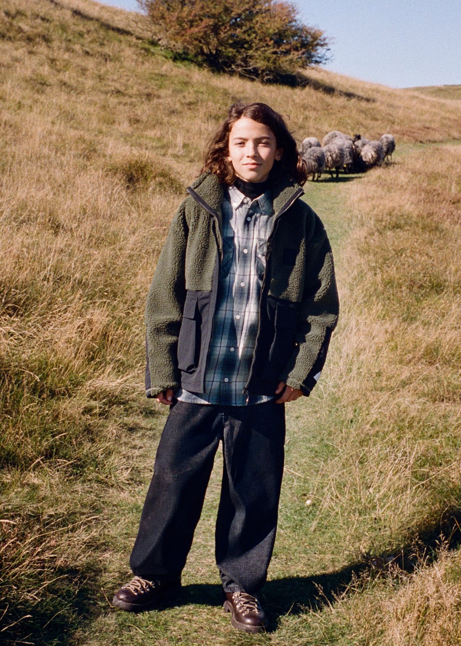 A person standing outdoors on a grassy hillside wearing a dark olive green hooded jacket over a blue and white plaid shirt, dark pants, and brown shoes. Sheep are visible in the background on the hill under a clear blue sky.