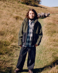 A person standing outdoors on a grassy hillside wearing a dark olive green hooded jacket over a blue and white plaid shirt, dark pants, and brown shoes. Sheep are visible in the background on the hill under a clear blue sky.