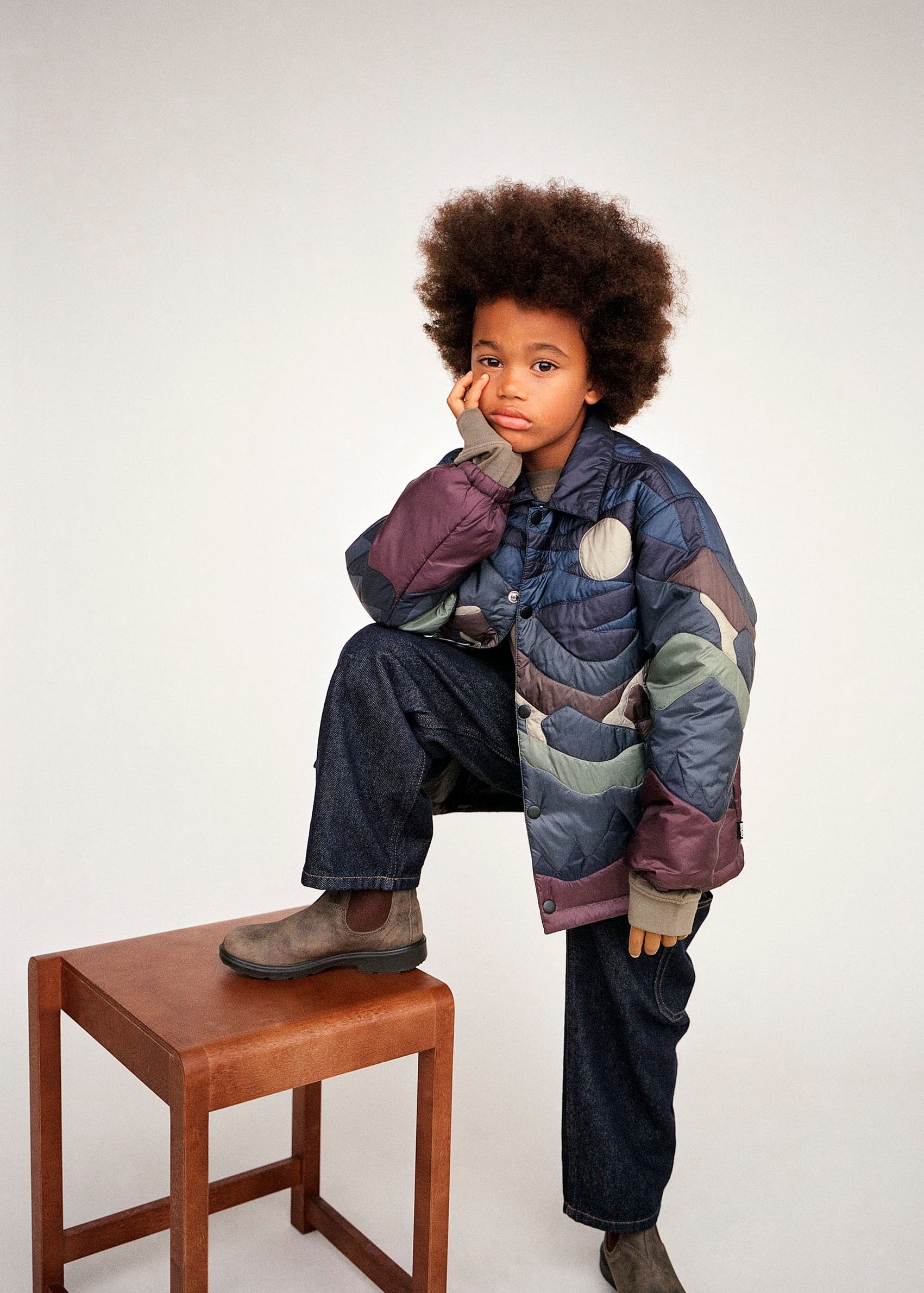 Child wearing a colorful puffer jacket sitting on a wooden stool against a white background