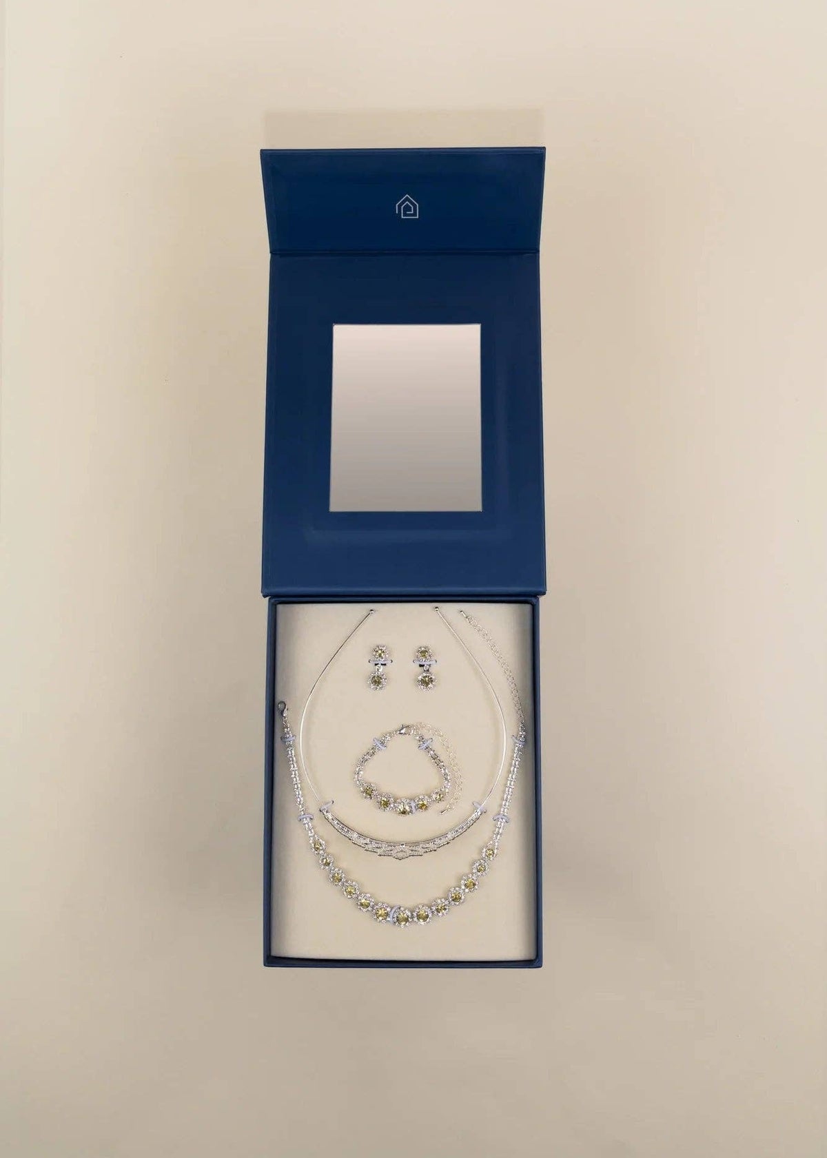 A jewelry set displayed inside an open dark blue presentation box on a light beige background. The set includes a necklace, bracelet, and matching earrings with clear rhinestone-like embellishments on silver-toned chains.