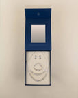 A jewelry set displayed inside an open dark blue presentation box on a light beige background. The set includes a necklace, bracelet, and matching earrings with clear rhinestone-like embellishments on silver-toned chains.
