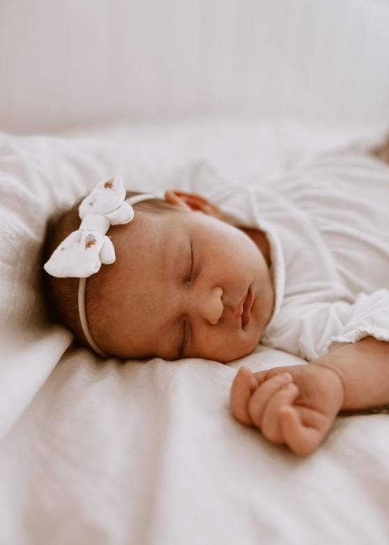 Newborn baby sleeping peacefully with a white bow headband on a soft surface.