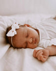 Newborn baby sleeping peacefully with a white bow headband on a soft surface.