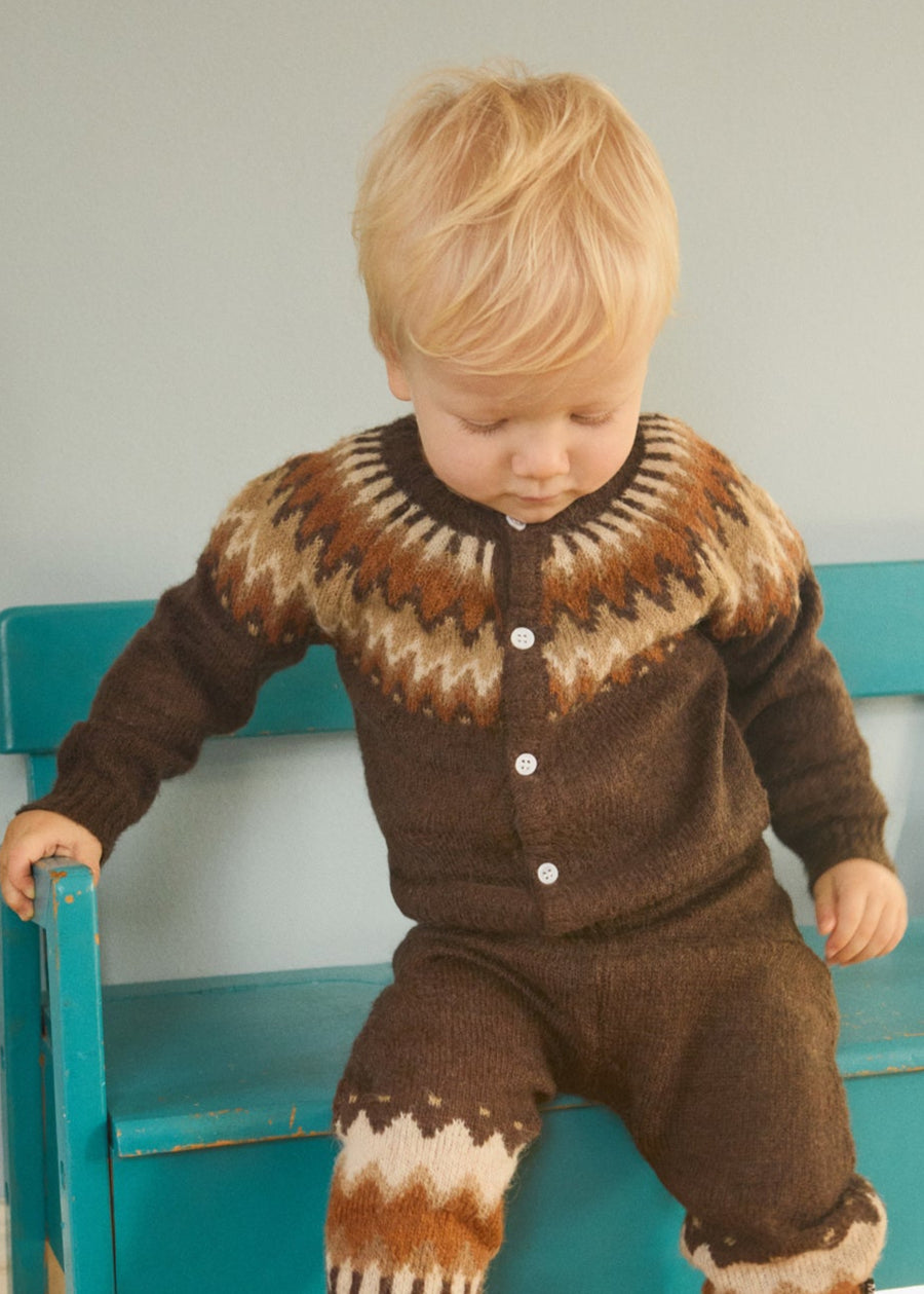 Child wearing a brown patterned outfit standing on a wooden floor with a teal bench.