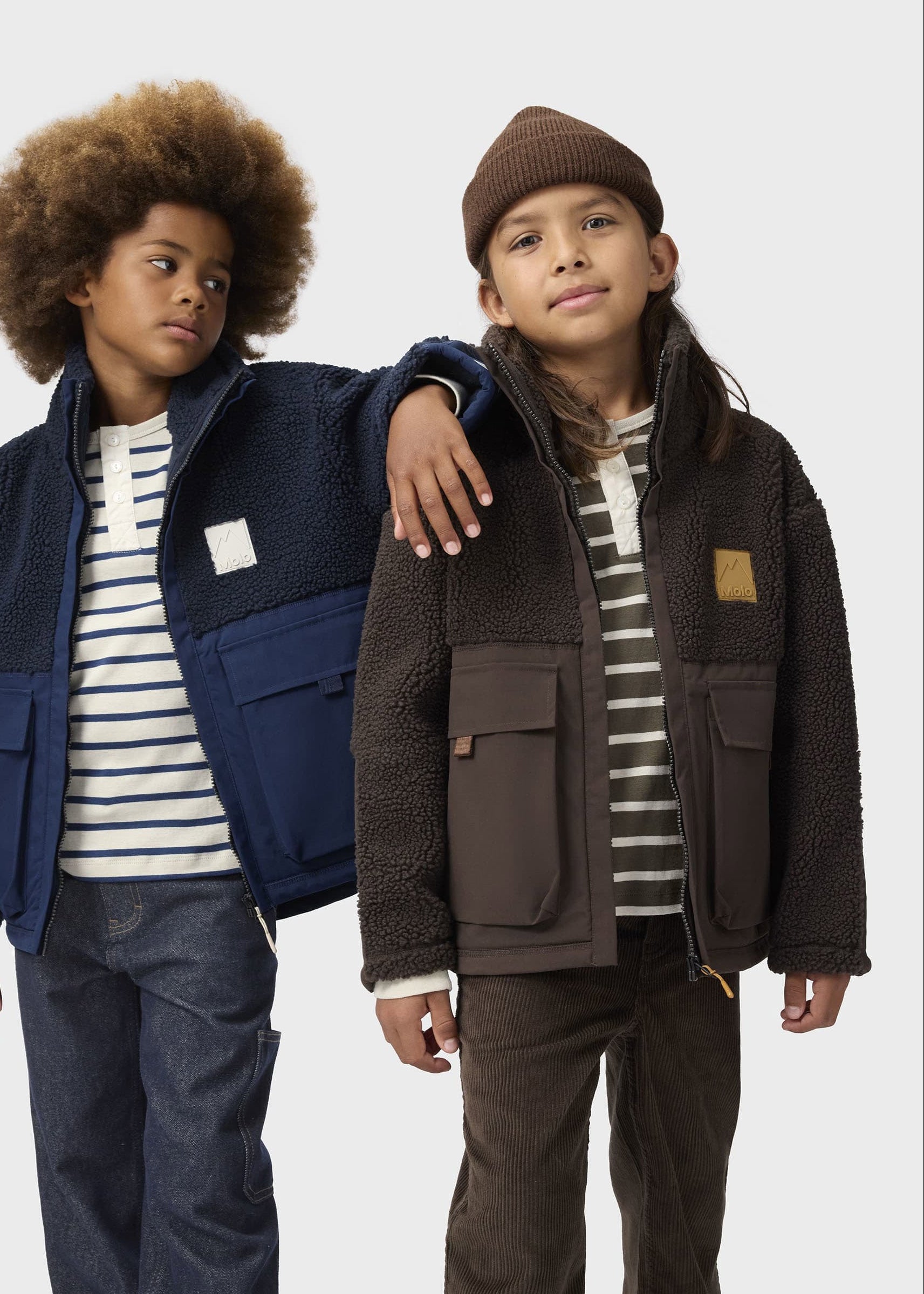 Two children modeling winter jackets against a light gray studio background. The child on the left wears a navy and blue jacket over a white and navy striped shirt with dark jeans.