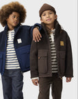 Two children modeling winter jackets against a light gray studio background. The child on the left wears a navy and blue jacket over a white and navy striped shirt with dark jeans.