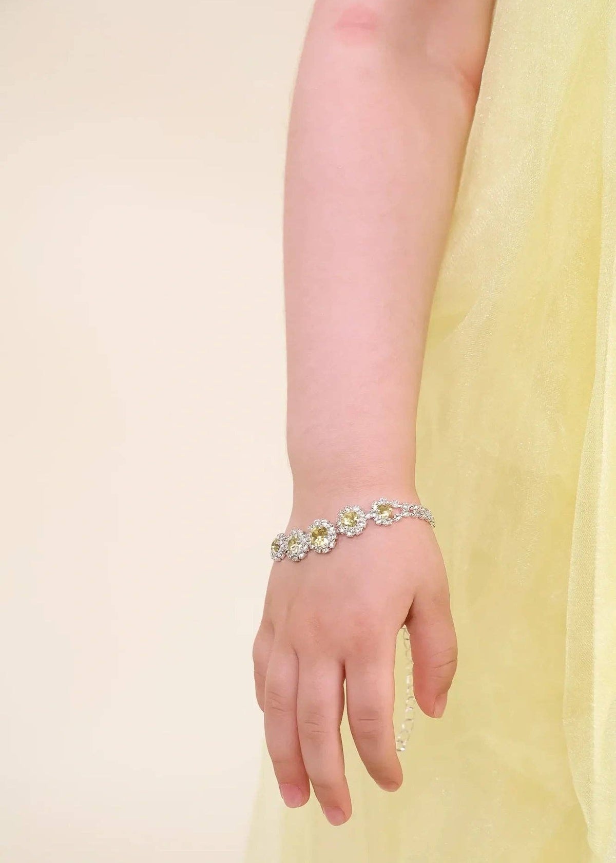 A close-up of a child's hand and wrist wearing a delicate silver-toned bracelet adorned with small white and yellow floral details, set against a soft cream background and a pale yellow dress.