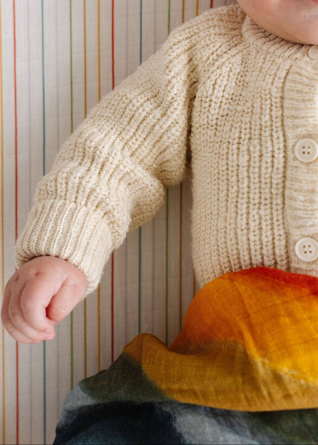 Child wearing a beige knitted sweater with colorful pants against a striped background