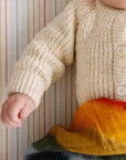 Child wearing a beige knitted sweater with colorful pants against a striped background