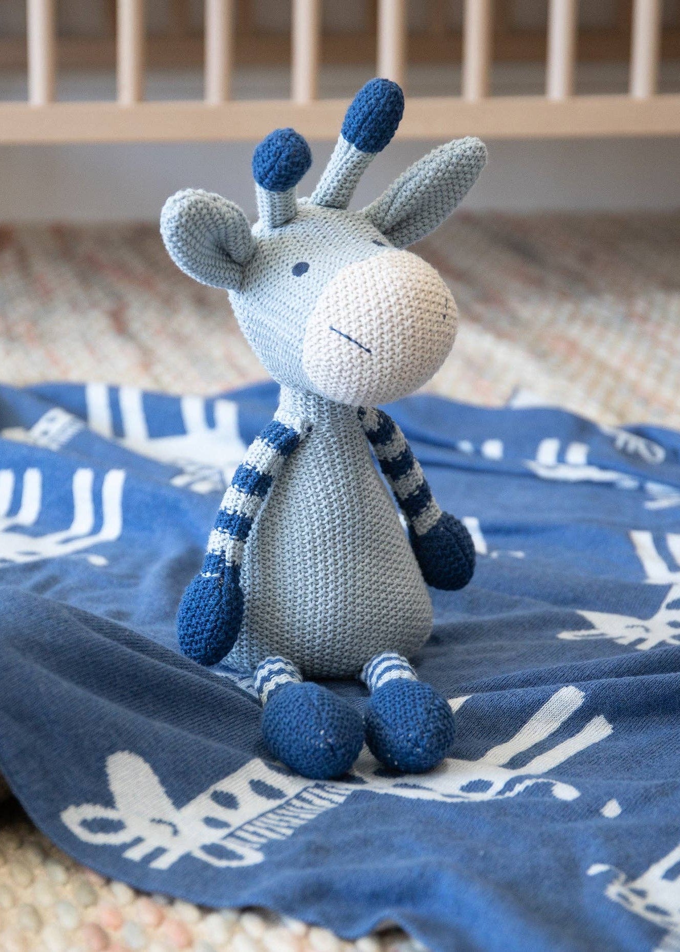 A crocheted giraffe plush toy in light gray and navy blue sits on a blue blanket with white giraffe patterns, placed near a crib.