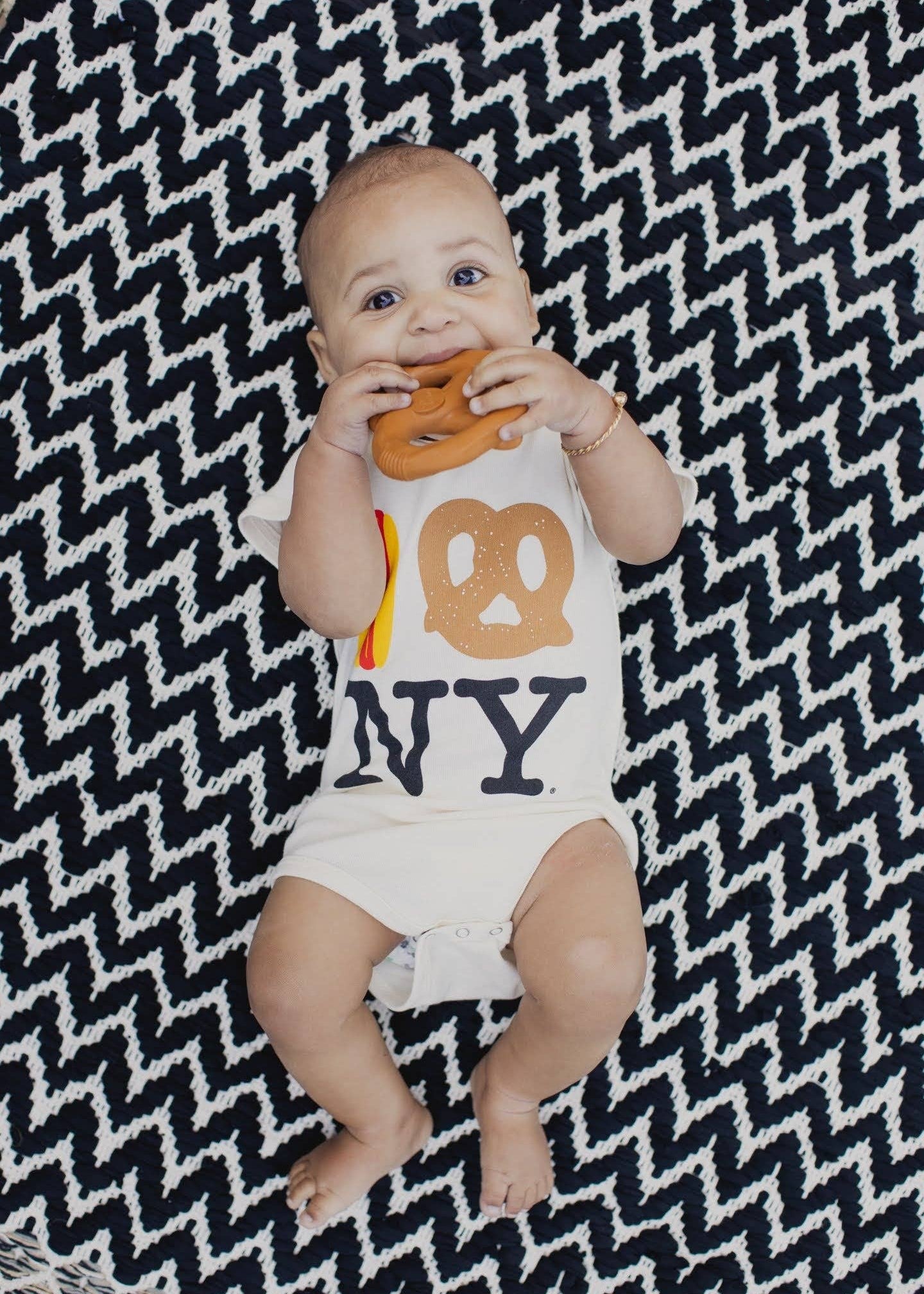 A baby lying on a black and white chevron blanket, wearing a white bodysuit with a pretzel and 'NY' graphic, while holding a pretzel toy.