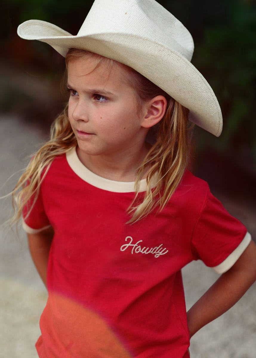 Young girl wearing a white cowboy hat and red 'Howdy' shirt outdoors.