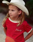 Young girl wearing a white cowboy hat and red 'Howdy' shirt outdoors.