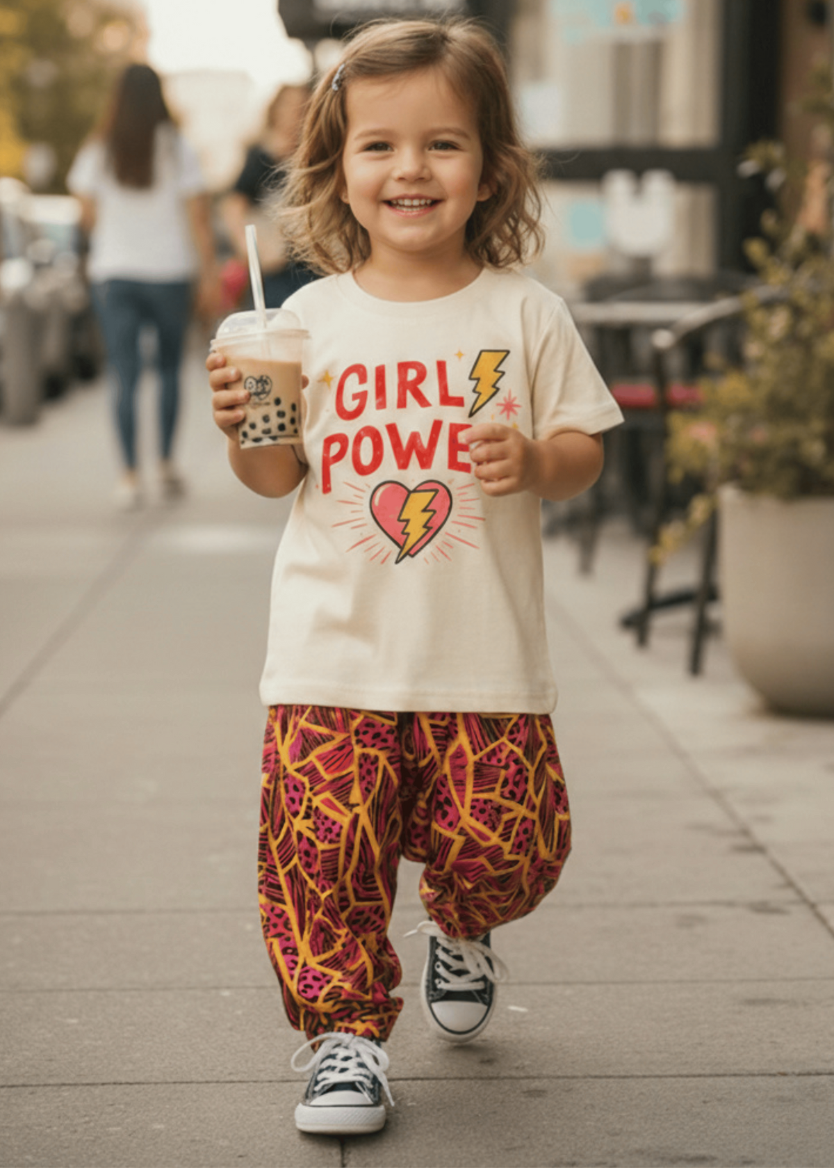 A young child walking on a city sidewalk wearing a cream graphic T-shirt that reads 'GIRL POWER' with a red and yellow heart/lightning design, paired with loose red and yellow patterned pants and black-and-white sneakers, while holding a drink.