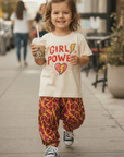 A young child walking on a city sidewalk wearing a cream graphic T-shirt that reads 'GIRL POWER' with a red and yellow heart/lightning design, paired with loose red and yellow patterned pants and black-and-white sneakers, while holding a drink.