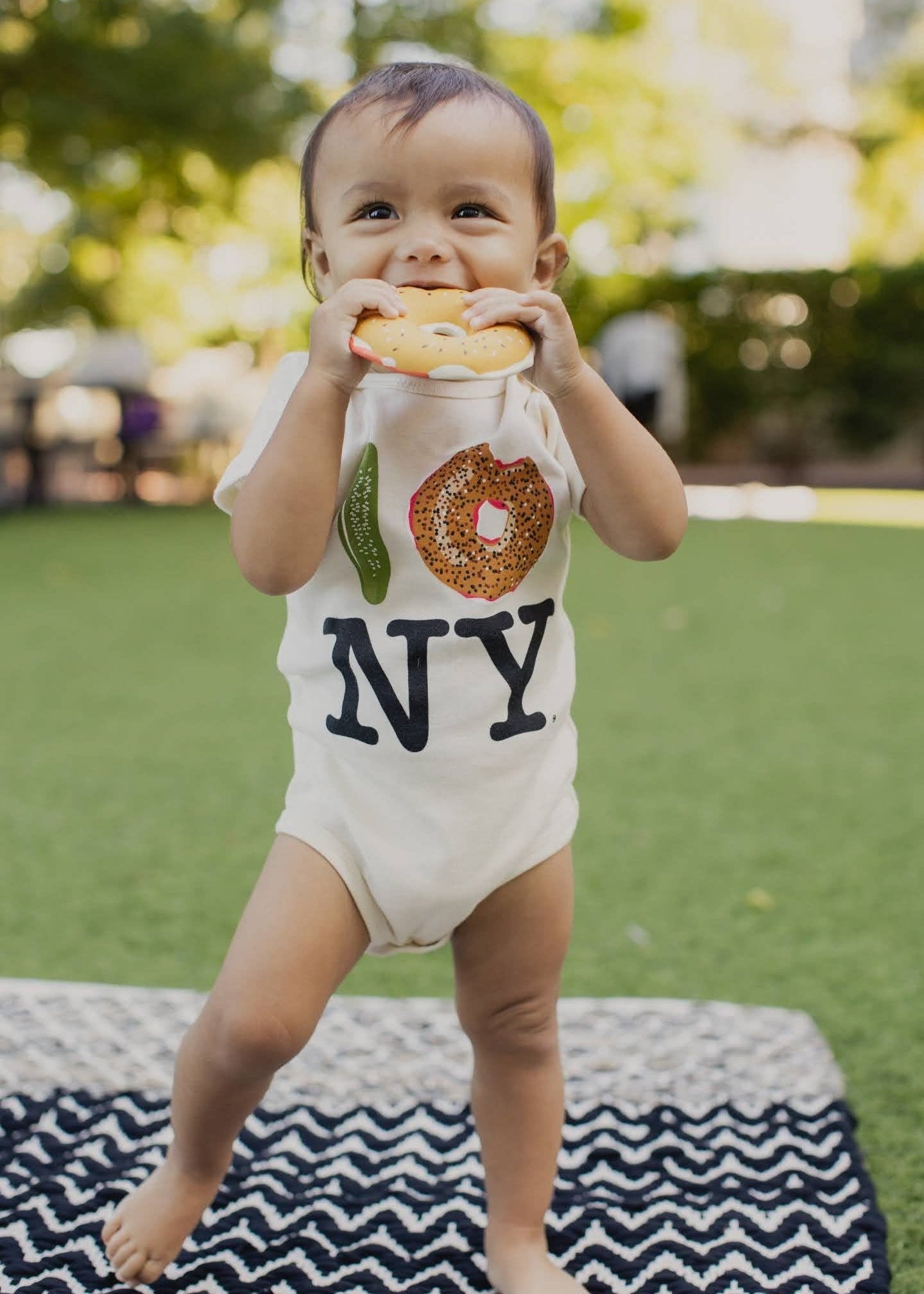 A baby standing outdoors on a black and white chevron blanket, wearing a cream bodysuit with a New York City skyline graphic and 'NY' text, while holding a bagel to their mouth.