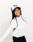 Child wearing a white long-sleeve shirt and black pants with a blue cap on a white background