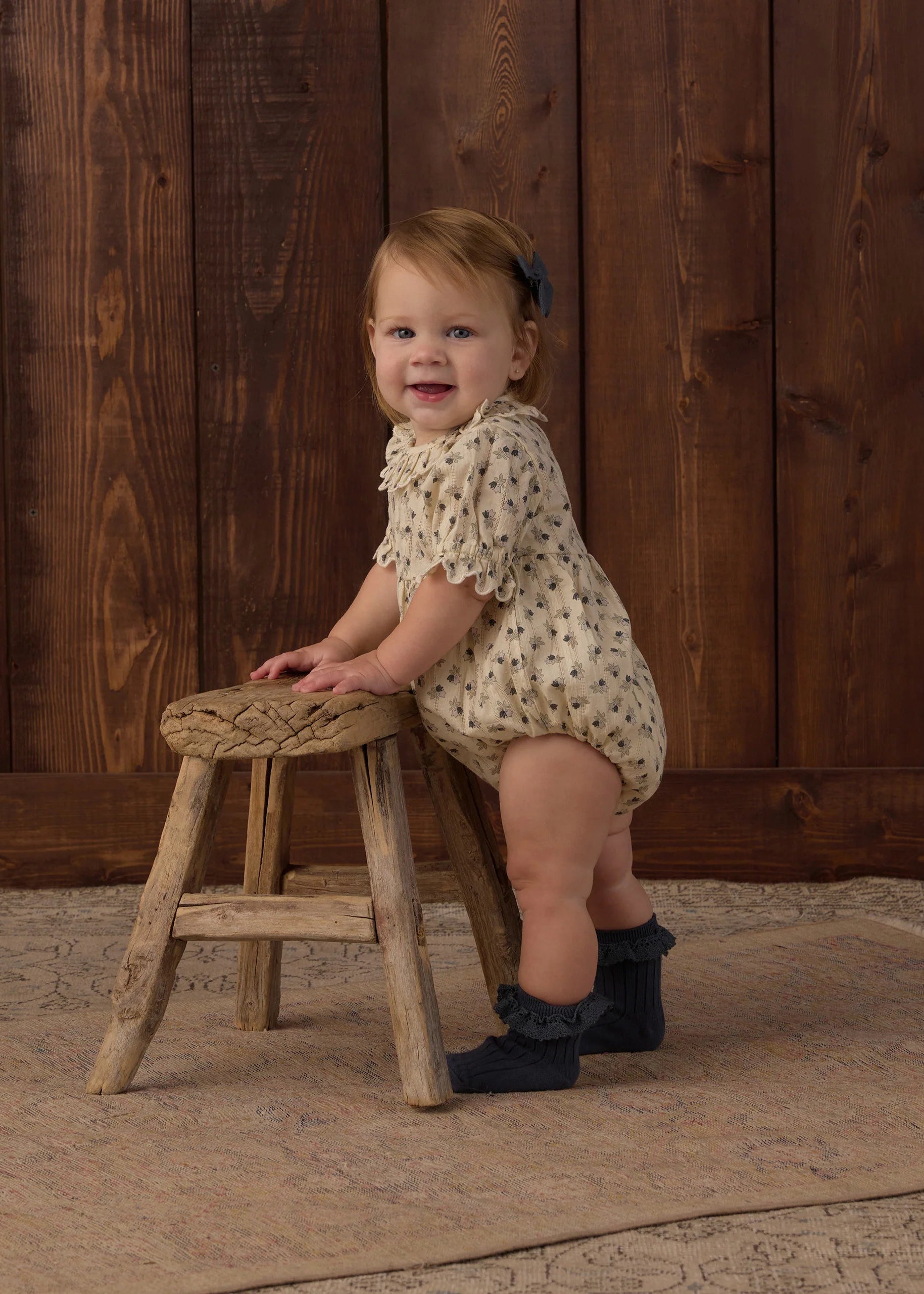 A smiling toddler stands on a rustic wooden stool against a dark wood plank wall, wearing a cream romper with small dark floral print and dark ruffled socks.