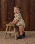 A smiling toddler stands on a rustic wooden stool against a dark wood plank wall, wearing a cream romper with small dark floral print and dark ruffled socks.