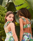 Two children wearing floral swimsuits standing in front of banana trees and large leaves.