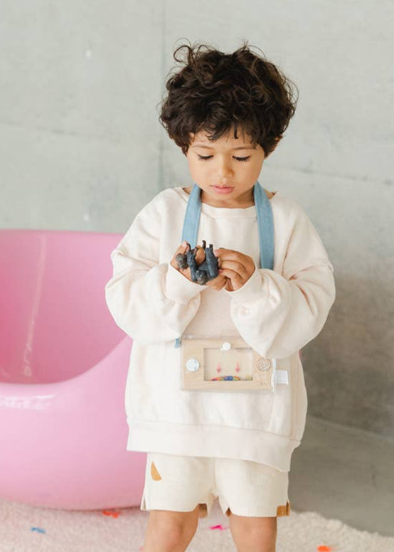 Child in a white outfit standing on a colorful rug with a pink chair in the background