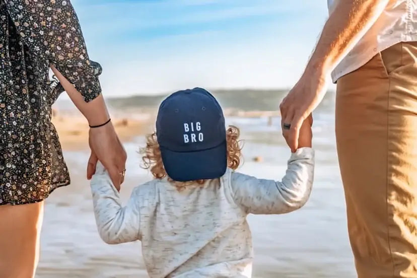 Child wearing a 'BIG BRO' cap on a beach with two adults holding hands