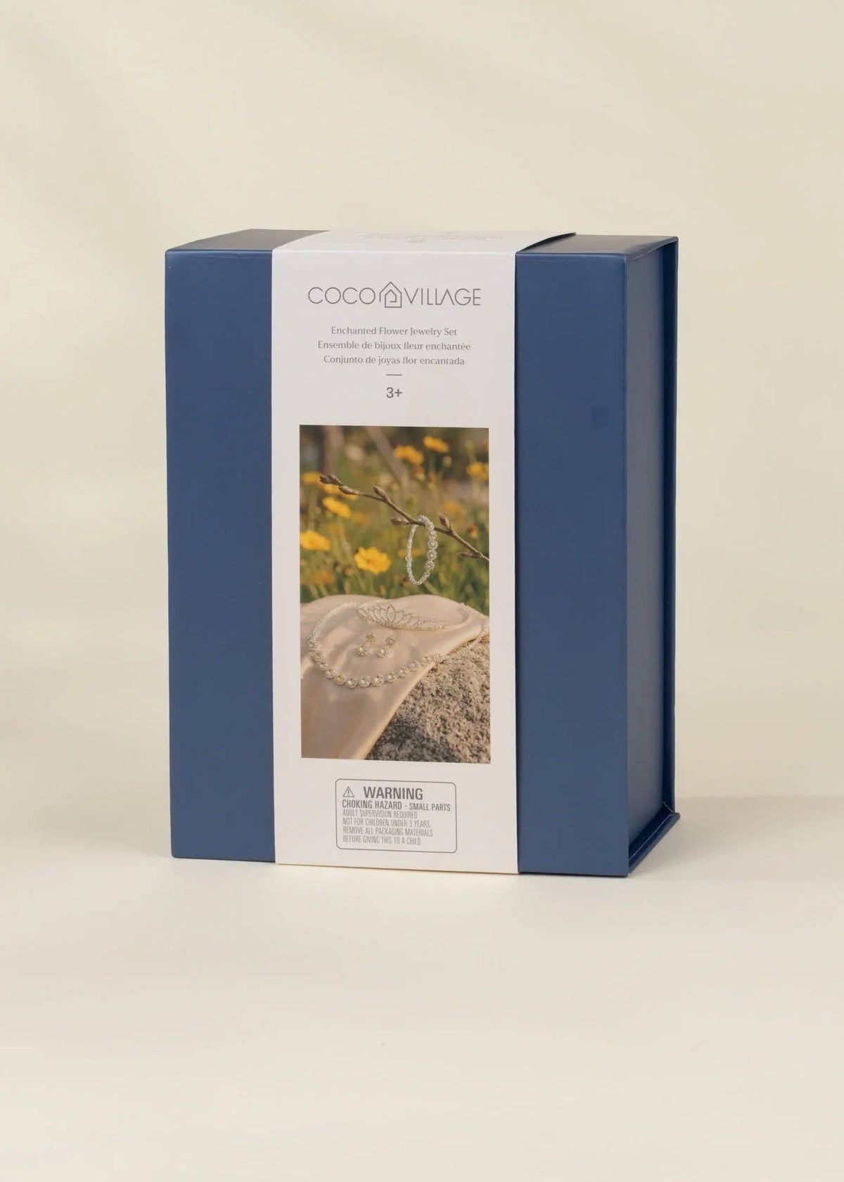 COCO VILAGE brand product box in navy blue and white packaging, featuring a photo of a beaded necklace on a stone with yellow flowers, set against a warm beige background.
