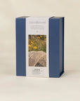 COCO VILAGE brand product box in navy blue and white packaging, featuring a photo of a beaded necklace on a stone with yellow flowers, set against a warm beige background.