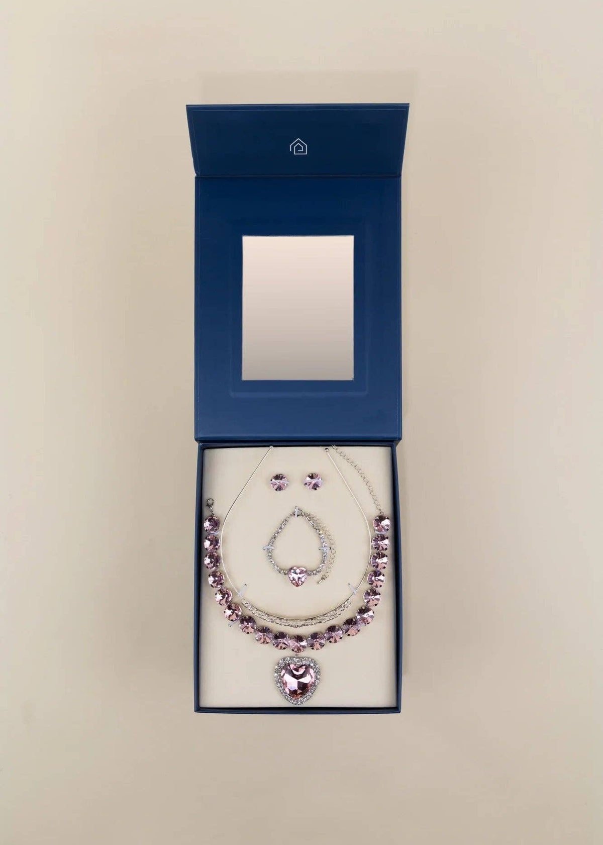 A silver-toned necklace and matching earrings set with light purple gemstones, presented in an open dark blue jewelry box with a mirror inside, set against a plain beige background.