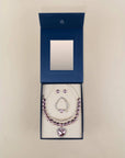 A silver-toned necklace and matching earrings set with light purple gemstones, presented in an open dark blue jewelry box with a mirror inside, set against a plain beige background.